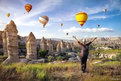 A hiker raises their arms in celebration on a rocky ridge surrounded by fairy chimneys in Cappadocia, Turkey, with colorful hot air balloons filling the sky above the ancient landscape.