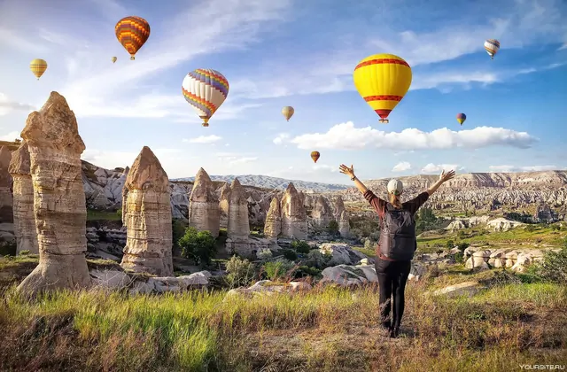 Un escursionista alza le braccia in celebrazione su una cresta rocciosa circondata da camini di fate in Cappadocia, Turchia, con variopinte mongolfiere che riempiono il cielo sopra l'antico paesaggio.
