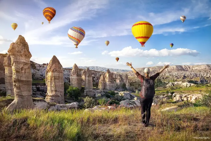 Un escursionista alza le braccia in celebrazione su una cresta rocciosa circondata da camini di fate in Cappadocia, Turchia, con variopinte mongolfiere che riempiono il cielo sopra l'antico paesaggio.