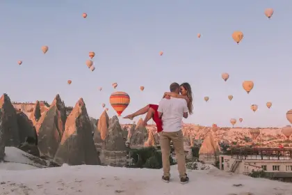 A man lifts his partner for a romantic photo at dawn in Cappadocia, Turkey, with the glowing sky full of ascending hot air balloons forming an unforgettable backdrop.