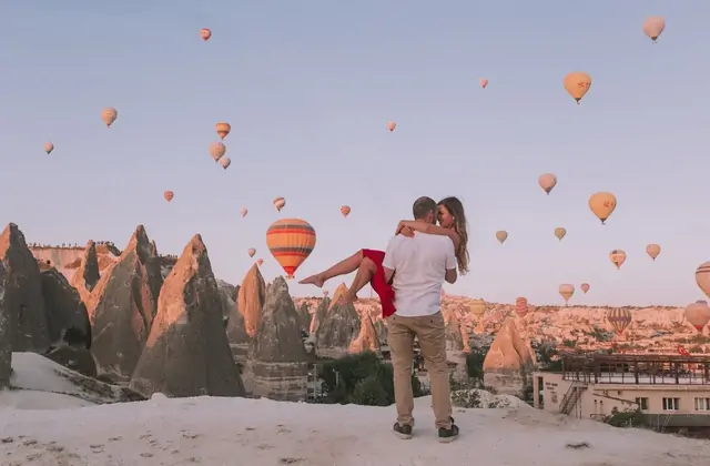 Un uomo solleva la sua partner per una foto romantica all'alba in Cappadocia, Turchia, con il cielo luminoso pieno di mongolfiere in ascesa che formano uno sfondo indimenticabile.
