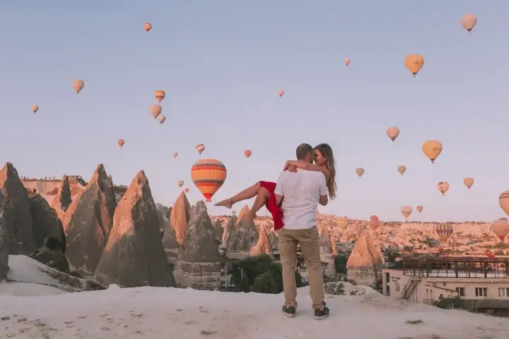 Un uomo solleva la sua partner per una foto romantica all'alba in Cappadocia, Turchia, con il cielo luminoso pieno di mongolfiere in ascesa che formano uno sfondo indimenticabile.