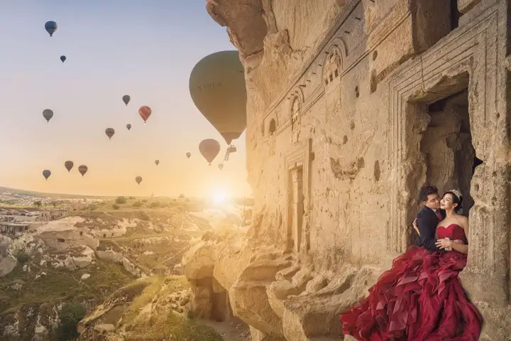 Una coppia posa per un ritratto accanto a un'antica incisione rupestre in Cappadocia, Turchia, con mongolfiere visibili nel cielo sopra la scogliera di tufo intagliata dietro di loro.