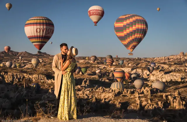 Una coppia condivide un tenero bacio in Cappadocia, Turchia, con un cielo pieno di mongolfiere multicolori morbidamente sfocate sullo sfondo, creando una scena per eccellenza romantica.