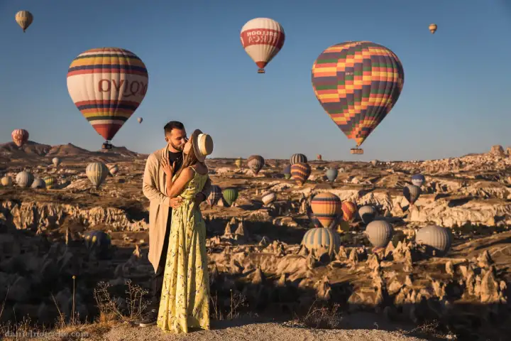 Una pareja comparte un tierno beso en Capadocia, Turquía, con un cielo lleno de globos aerostáticos multicolores suavemente desenfocados en el fondo, creando una escena quintaesencialmente romántica.