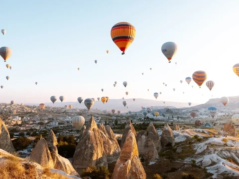 Al crepuscolo, sulla terrazza di un hotel in grotta della Cappadocia, una coppia osserva le ultime mongolfiere del giorno passare davanti al cielo violetto sopra le silhouette rocciose della valle.