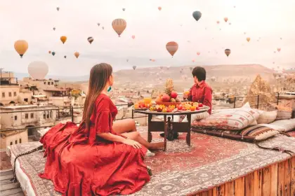 A traditional Turkish breakfast spread is laid on a rooftop terrace in Cappadocia, Turkey, framing a sunrise sky where hot air balloons rise above the ancient stone town below.