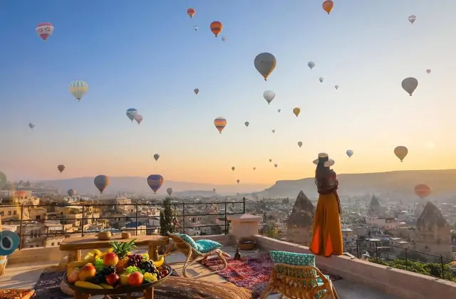 Una donna si appoggia alla balaustra in pietra di una terrazza di hotel grotta della Cappadocia, Turchia, guardando verso l'alto un cielo affollato di mongolfiere nella dorata luce mattutina.