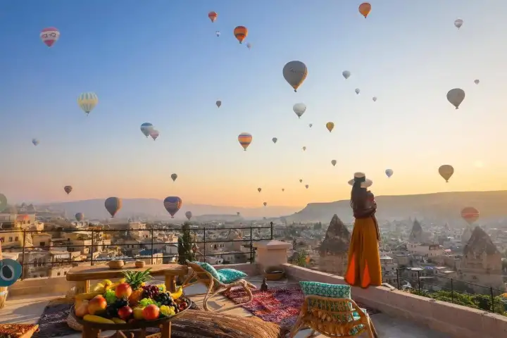 Una mujer se apoya en la balaustrada de piedra de una terraza de hotel cueva de Capadocia, Turquía, mirando hacia arriba un cielo repleto de globos aerostáticos en la dorada luz matinal.