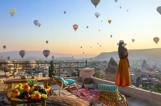 Standing at a rooftop viewpoint in Cappadocia, Turkey, a woman watches the pre-dawn sky fill with the glowing envelopes of dozens of hot air balloons beginning their morning ascent.
