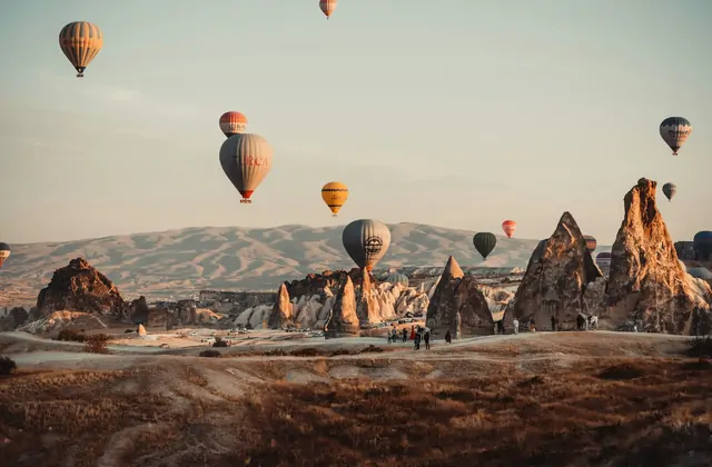 Dramáticas agujas de roca se elevan del paisaje de Capadocia, Turquía, mientras los globos aerostáticos se entrelazan entre las altas formaciones en una escena que mezcla perfectamente la maravilla natural con la aventura aérea.