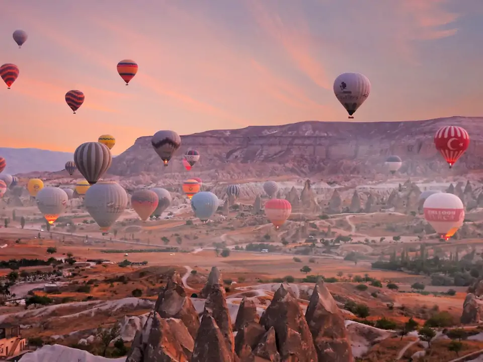Davanti a un cielo rosato dell’alba, diverse mongolfiere scivolano sopra una valle di camini delle fate in Cappadocia, Turchia, mentre la morbida luce rosa tinge i coni vulcanici di calde sfumature pesca.