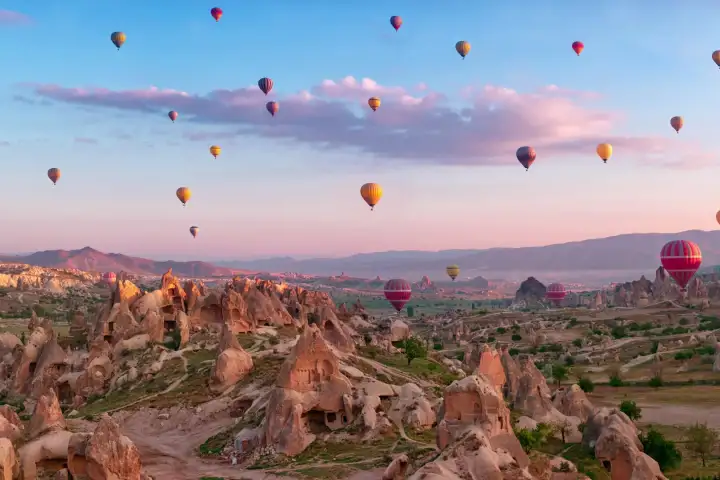 A pink sky at dawn envelops the valleys of Cappadocia, Turkey, as hot air balloons in every color rise above the pale rock ridges and silent village rooftops below.