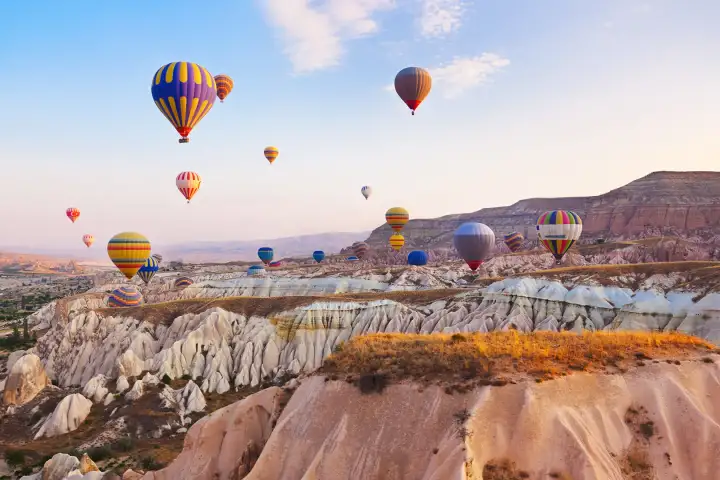 Rainbow-striped and multicolored hot air balloons sail above a white volcanic ridge landscape in Cappadocia, Turkey, the bright spectrum of colors vivid against the pale stone formations.