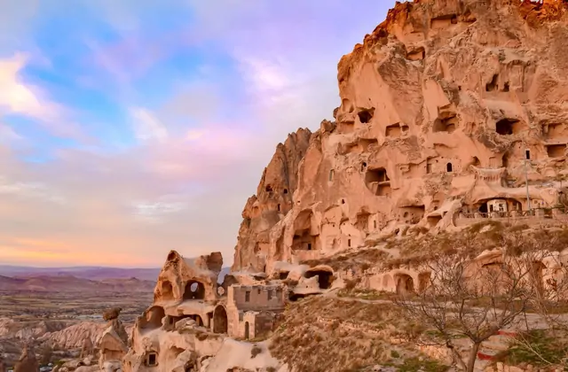 Las casas cueva excavadas en roca se agrupan a lo largo de una ladera de Capadocia al atardecer, Turquía, sus ventanas brillando ámbar mientras la última luz del sol esculpe profundas sombras en las fachadas de piedra volcánica tallada.