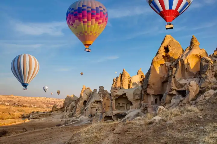 Hot air balloons drift directly overhead above the rock-cut cave houses of Cappadocia, Turkey, their colorful forms reflected in the windows of the ancient stone dwellings below.