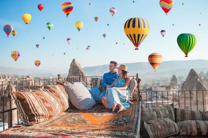 A couple portrait taken on a stone terrace in Cappadocia, Turkey, with a vibrant sky packed with dozens of vivid hot air balloons creating an extraordinary background of color and movement.