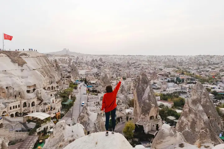 A traveler in a bright red jacket stands at the Göreme viewpoint in Cappadocia, Turkey, the vivid jacket contrasting with the grey and ochre of the cave-carved valley below.