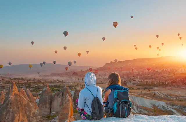 Due zaini in spalla si siedono a un belvedere al tramonto della Cappadocia, Turchia, le loro zaini accanto mentre guardano lo spettacolo di mongolfiere che derivano nel cielo arancione che si fa più profondo.