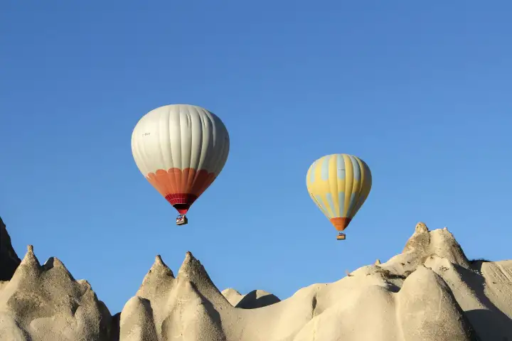 Two hot air balloons float side by side above the distinctive white volcanic rock formations of Cappadocia, Turkey, their colorful envelopes a vivid contrast against the pale stone landscape.