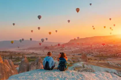 Two travelers perch on the edge of a cliff at sunrise in Cappadocia, Turkey, watching in silence as a procession of hot air balloons crosses the blazing horizon below their feet.