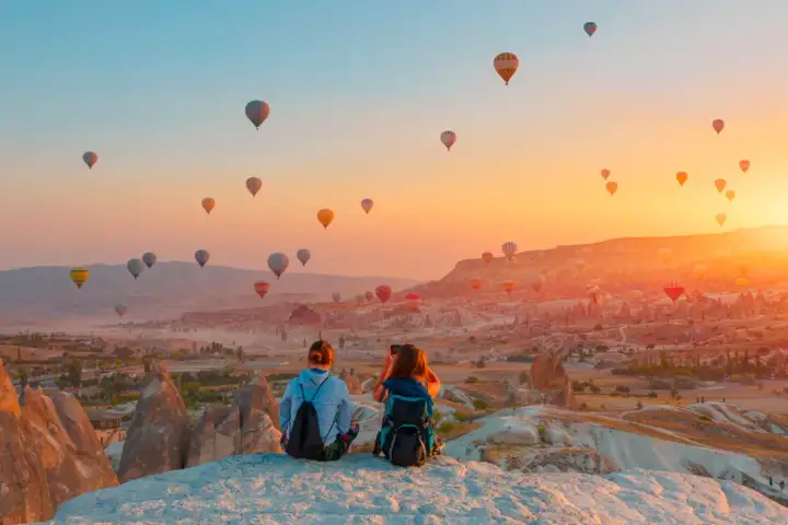 Due viaggiatori si appoggiano sul bordo di una scogliera all'alba in Cappadocia, Turchia, osservando in silenzio mentre una processione di mongolfiere attraversa l'ardente orizzonte sotto i loro piedi.