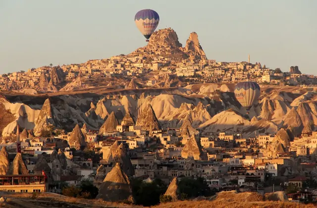 The towering volcanic mass of Uçhisar Castle rises above the Cappadocian landscape, Turkey, surrounded by fairy chimneys and framed by hot air balloons drifting through the clear morning air.