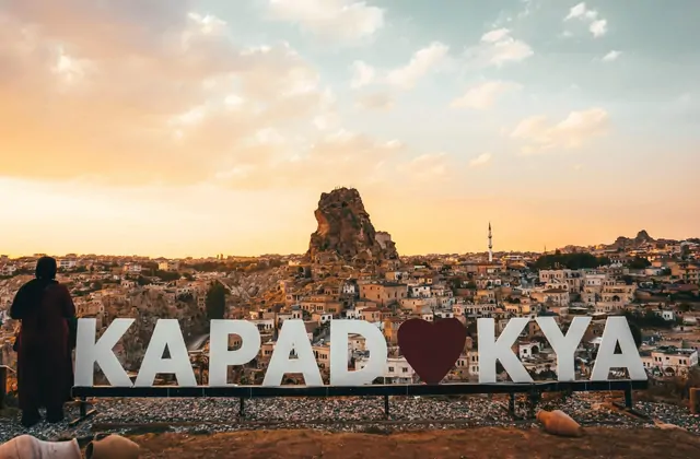 A wide panorama of Uçhisar town in Cappadocia, Turkey, features the iconic 'Kapadokya' sign in the foreground with the castle rock and cave-pocked hillside stretching across the frame.