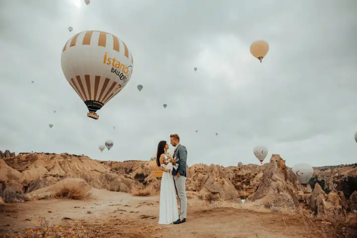 A wedding couple poses for a portrait in Cappadocia, Turkey, with a dramatic cloudy sky behind them and colorful hot air balloons breaking through the clouds above the volcanic landscape.