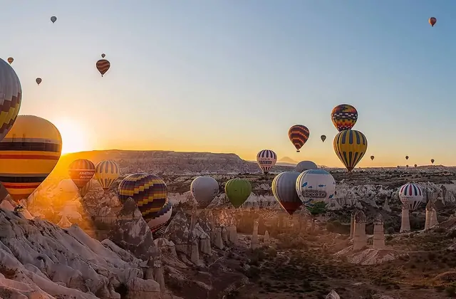 A sun flare bursts across a morning view over Göreme, Cappadocia, Turkey, with hot air balloons drifting through the lens flare and golden light above the ancient cave town.