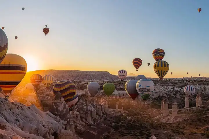 Un bagliore solare esplode su una vista mattutina su Göreme, Cappadocia, Turchia, con mongolfiere che derivano attraverso il riflesso dell'obiettivo e la luce dorata sopra l'antico borgo rupestre.