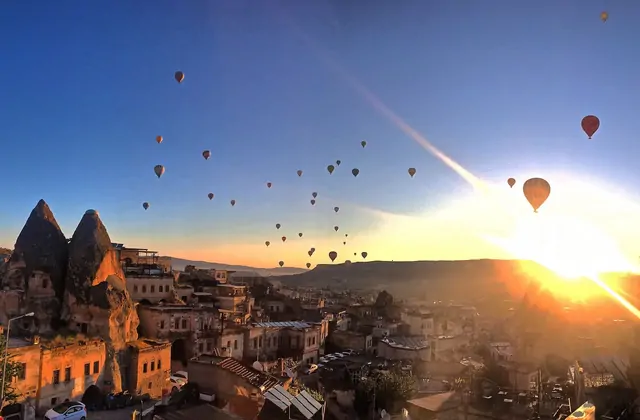 Sunrise breaks over a cave town in Cappadocia, Turkey, the warm light illuminating carved rooftops and minarets while a skyline of ascending hot air balloons rises behind the stone silhouettes.