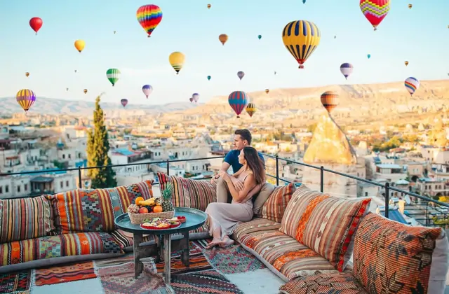 A person strikes a yoga-style pose on a Cappadocian rooftop at sunrise, Turkey, arms open wide as dozens of hot air balloons drift through the pink and gold sky behind them.