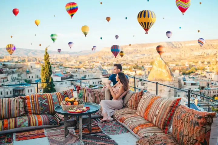 A person strikes a yoga-style pose on a Cappadocian rooftop at sunrise, Turkey, arms open wide as dozens of hot air balloons drift through the pink and gold sky behind them.