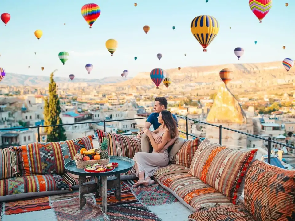 A person strikes a yoga-style pose on a Cappadocian rooftop at sunrise, Turkey, arms open wide as dozens of hot air balloons drift through the pink and gold sky behind them.