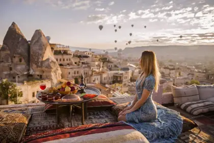 A woman sips morning tea on a terrace breakfast setting in Cappadocia, Turkey, while the sunrise sky behind her fills with colorful hot air balloons rising above the cave-carved valley.