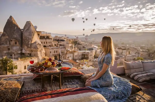 Una donna sorsa il tè mattutino in una colazione in terrazza in Cappadocia, Turchia, mentre il cielo all'alba dietro di lei si riempie di variopinte mongolfiere che si alzano sopra la valle rupestre.