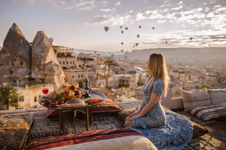 A woman sips morning tea on a terrace breakfast setting in Cappadocia, Turkey, while the sunrise sky behind her fills with colorful hot air balloons rising above the cave-carved valley.