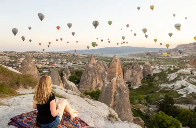 Una donna siede su un tradizionale tappeto turco a un punto panoramico all'aperto in Cappadocia, Turchia, guardando le mongolfiere che derivano serenamente sopra i famosi camini di fate nella valle davanti a lei.