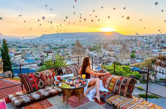 A woman enjoys a solo rooftop breakfast in Cappadocia, Turkey, the table set with Turkish morning foods while behind her the sunrise sky is alive with ascending hot air balloons.