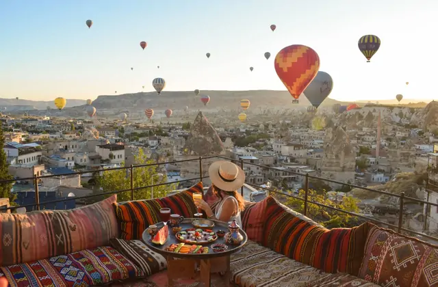 A woman wearing a wide-brimmed hat stands on a Cappadocian terrace, Turkey, looking toward the horizon where colorful hot air balloons dot the blue sky above the rock formations.