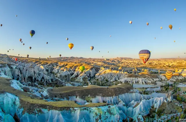 A panoramic view of White Valley in Cappadocia, Turkey, reveals the pale, chalky rock formations stretching into the distance while morning hot air balloons hover above the bleached canyon landscape.