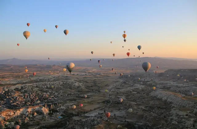 From high above, a wide aerial view of a Cappadocian valley, Turkey, shows a dense cloud of morning hot air balloons clustered together, their colors forming a vivid mosaic in the air.