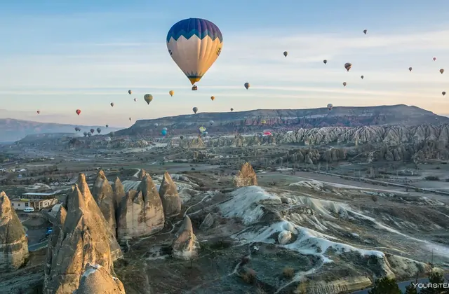 Una mongolfiera gigante domina l'inquadratura in una scena di valle ampia all'alba in Cappadocia, Turchia, il suo enorme involucro che si erge sopra le formazioni rocciose mentre il cielo si schiarisce intorno ad essa.