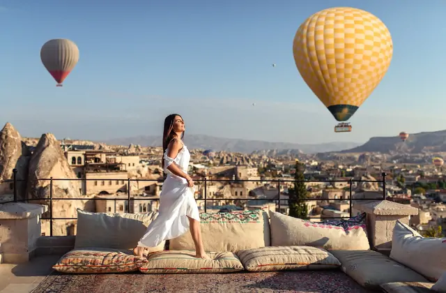 A woman in a flowing white dress stands on a carved stone terrace in Cappadocia, Turkey, facing the morning sky where colorful hot air balloons drift above the valley below.