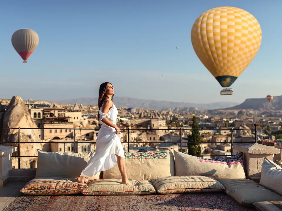 Una mujer con un vestido blanco vaporoso está de pie en una terraza de piedra tallada en Capadocia, Turquía, mirando al cielo de la mañana mientras coloridos globos aerostáticos flotan sobre el valle.