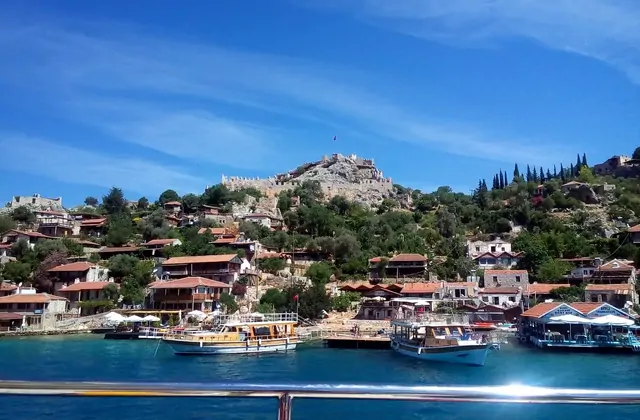 Barcos turísticos de madera están amarrados en el pequeño puerto de Kaleköy cerca de Kekova, provincia de Antalya, con el pueblo de piedra en terrazas y los muros en ruinas del Castillo de Simena elevándose contra un cielo azul intenso, fotografiados desde la barandilla de un barco.