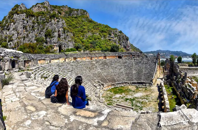 Tres visitantes se sientan en los desgastados asientos de piedra del antiguo teatro romano de Myra, provincia de Antalya, mirando a través de la amplia cavea semicircular, con fachadas de tumbas licianas talladas en la roca visibles en el acantilado gris que se eleva directamente detrás del escenari