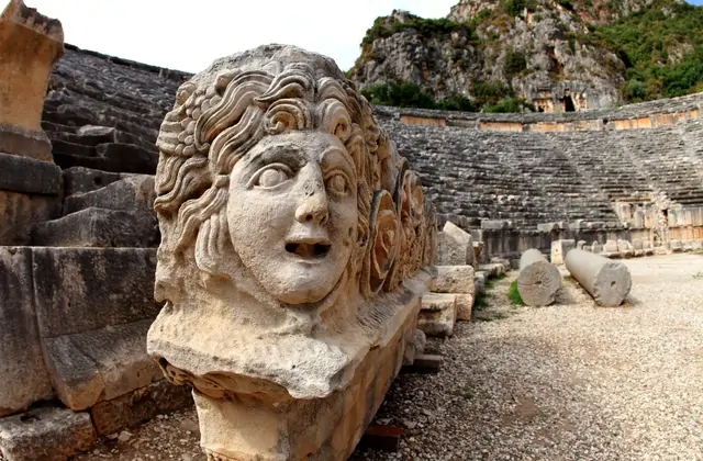 Una gran escultura de piedra con máscara de Medusa tallada se encuentra en primer plano del antiguo teatro romano de Myra en la provincia de Antalya, su cabello rizado salvaje y boca abierta representados en dramático relieve, con las filas de asientos de piedra curvos y el acantilado licio elevándo