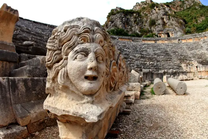 Una gran escultura de piedra con máscara de Medusa tallada se encuentra en primer plano del antiguo teatro romano de Myra en la provincia de Antalya, su cabello rizado salvaje y boca abierta representados en dramático relieve, con las filas de asientos de piedra curvos y el acantilado licio elevándo